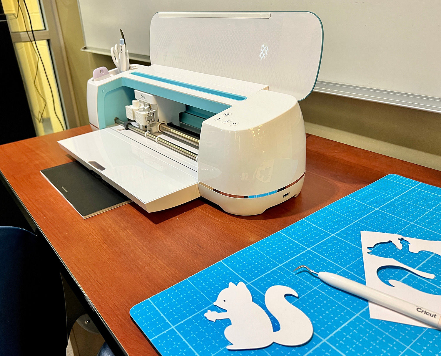 a freshly-cut cardstock squirrel sits on a cutting mat in front of the Cricut Maker, a free-to-use machine located in the Student Multimedia Studio